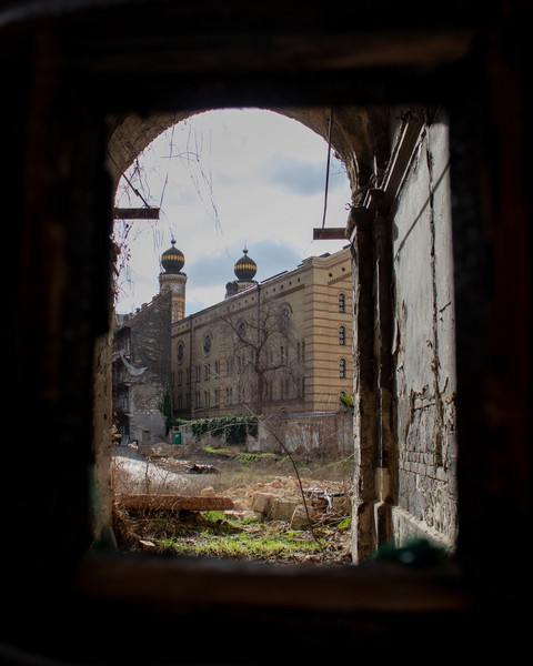 Durch einen baufälligen Torbogen sieht man im Vordergrund eine Brache und dahinter die Synagoge mit ihren zwei schmuckvollen Türmen.