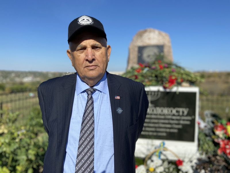 An elderly man in a suit and cap stands in front of a memorial stone decorated with flowers and a menorah.