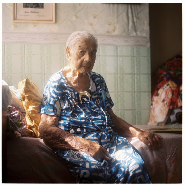 An old woman sits on the edge of a bed and looks seriously into the camera.