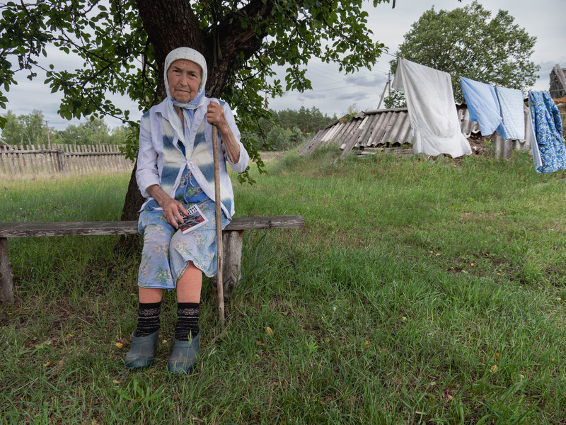 An old woman with a headscarf, weather-beaten skin, and a walking stick sits on a simple wooden bench in front of a tree; next to her, laundry on a line blows in the wind.